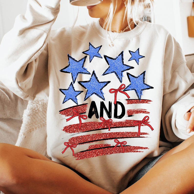 Pode incluir: Camisola branca com um design de bandeira americana vermelha, branca e azul. A bandeira tem listras vermelhas, estrelas brancas e la&ccedil;os vermelhos. A palavra "AND" est&aacute; escrita em letras pretas na bandeira.