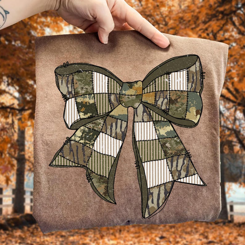 May include: A brown fabric with a large bow design. The bow features a patchwork pattern with camouflage and white striped sections. The bow is in shades of green, brown, and tan. The background is blurred, with fall foliage.
