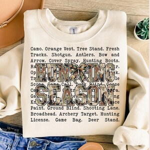 May include: A cream-colored sweatshirt with the words "HUNTING SEASON" in a camouflage pattern. The text is surrounded by a list of hunting-related terms. A decorative bow is between the words.