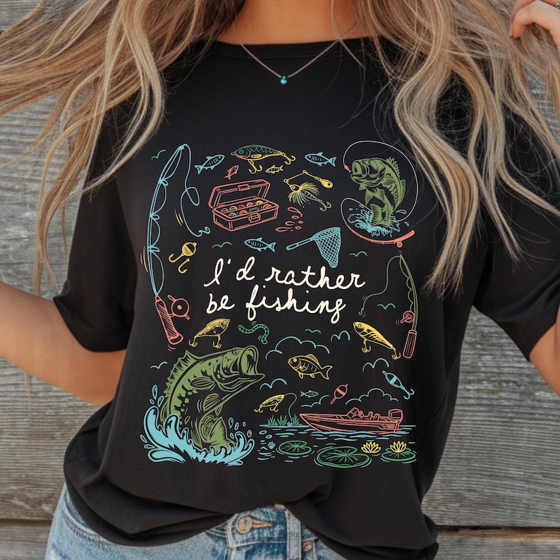 May include: Black t-shirt with a colorful fishing-themed graphic. The design includes fishing rods, fish, a tackle box, and the text "I'd rather be fishing."