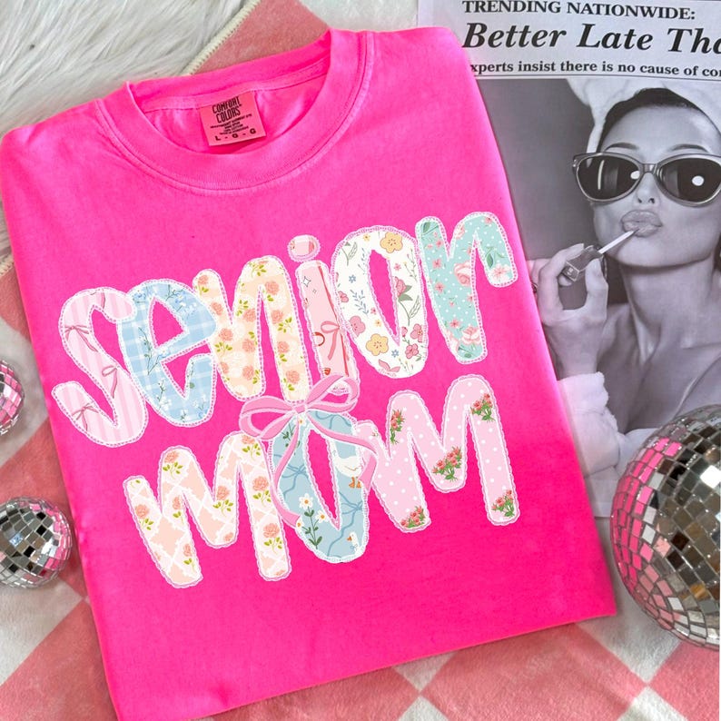 Puede incluir: Una camiseta rosa brillante con las palabras "Senior Mom" en un dise&ntilde;o de patchwork. Las letras est&aacute;n decoradas con telas florales y estampadas, y un lazo rosa est&aacute; centrado debajo de la palabra "Senior". La camiseta se muestra sobre una superficie a cuadros rosa y blanca.