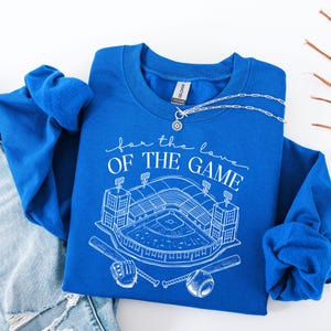 Op de afbeelding: Koningsblauw sweatshirt met een witte afbeelding van een honkbalstadion, knuppels en een honkbal. De tekst "for the love of the game" staat boven het stadionontwerp. Een zilveren ketting ligt over het sweatshirt.