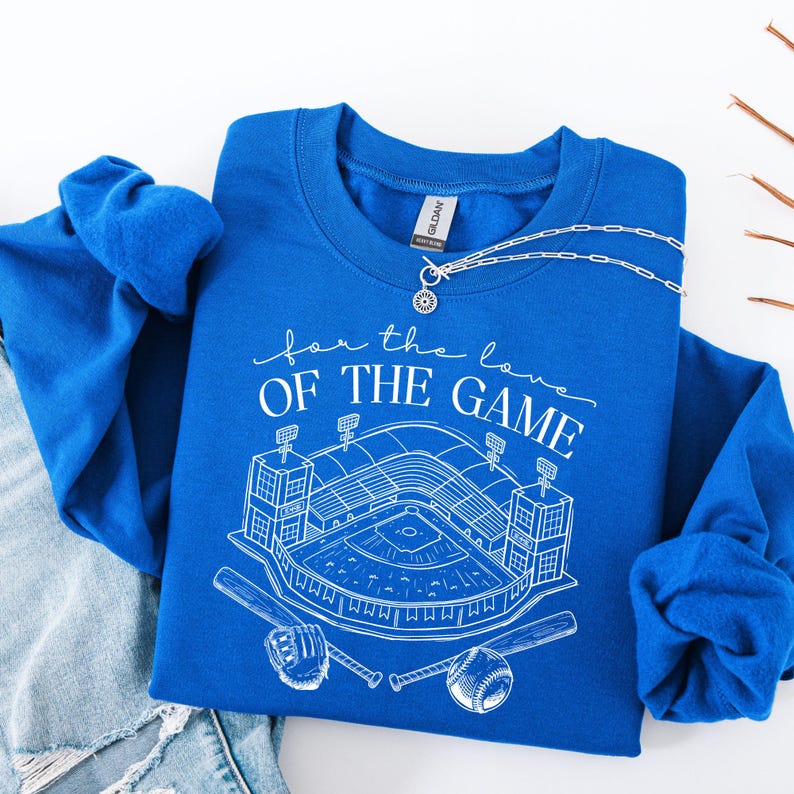 Op de afbeelding: Koningsblauw sweatshirt met een witte afbeelding van een honkbalstadion, knuppels en een honkbal. De tekst "for the love of the game" staat boven het stadionontwerp. Een zilveren ketting ligt over het sweatshirt.