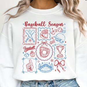 May include: White sweatshirt with a baseball-themed graphic in red and blue. The design includes baseball bats, a glove, a stadium, and the text "Baseball Season". The sweatshirt has a crew neck.