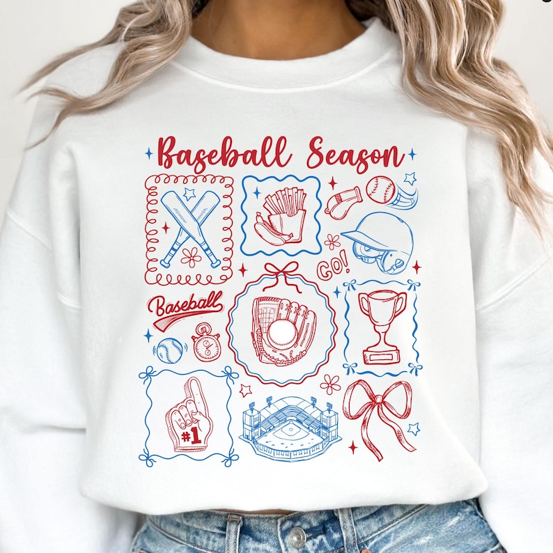 May include: White sweatshirt with a baseball-themed graphic in red and blue. The design includes baseball bats, a glove, a stadium, and the text "Baseball Season". The sweatshirt has a crew neck.