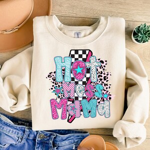 May include: A cream-colored sweatshirt with a graphic design that says "Hot Mess Mama" in pink, blue, and black letters. The graphic is surrounded by a checkered pattern, leopard print, and stars.