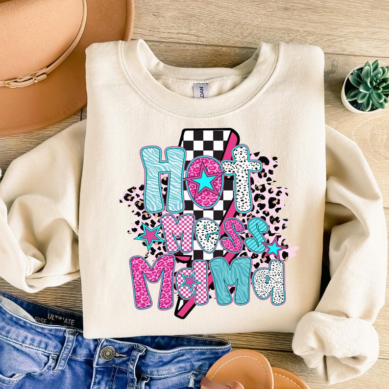 May include: A cream-colored sweatshirt with a graphic design that says "Hot Mess Mama" in pink, blue, and black letters. The graphic is surrounded by a checkered pattern, leopard print, and stars.
