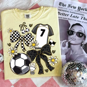 May include: A light yellow t-shirt with a soccer-themed design. The design includes a soccer ball, daisies, bows, and a jersey with the number 7. A newspaper with a black and white photo is in the background.