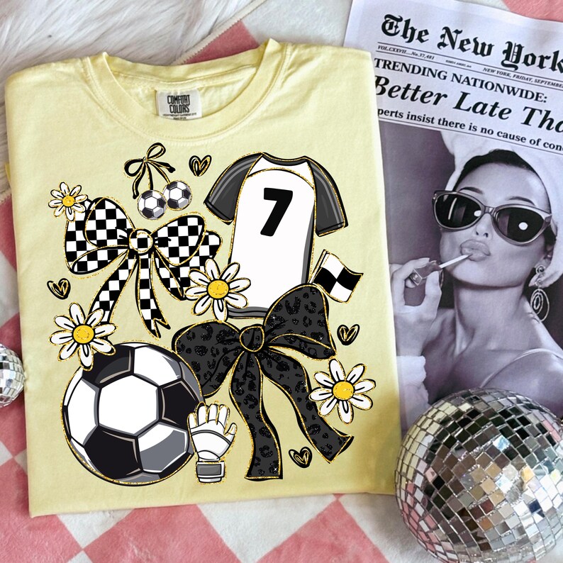 May include: A light yellow t-shirt with a soccer-themed design. The design includes a soccer ball, daisies, bows, and a jersey with the number 7. A newspaper with a black and white photo is in the background.