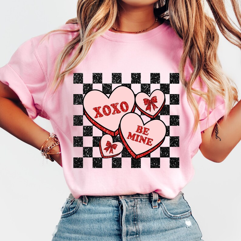 May include: Pink t-shirt with a black and white checkered pattern and three red glitter hearts. The hearts have the text "XOXO", "BE MINE", and a bow.