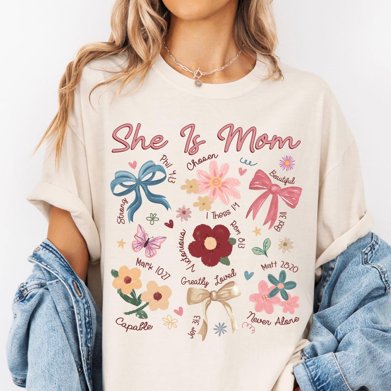 K&ouml;nnte beinhalten: Ein beiges T-Shirt mit dem Text "She Is Mom" in rosa Schrift. Das Shirt zeigt ein florales Design mit Schleifen, Schmetterlingen und Bibelversen.