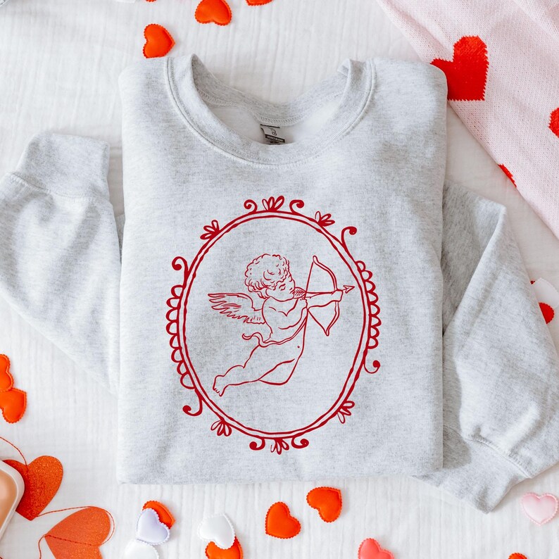 Pu&ograve; includere: Una felpa grigio chiaro con un disegno rosso incorniciato di Cupido con arco e freccia. L'illustrazione di Cupido &egrave; all'interno di una cornice ovale con decorazioni decorative. La felpa &egrave; piegata su una superficie bianca con decorazioni a cuore rosse.