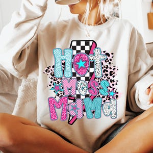 May include: A white sweatshirt with a graphic design featuring the text "Hot Mess Mama" in pink, blue, and black lettering with a checkered pattern and animal print accents.