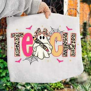 May include: White tote bag with the word "TEACH" in pink and tan letters. The letters "T" and "H" have a leopard print design. A ghost wearing a bow and holding a pencil is in the center. Pink bats and spiderwebs are also featured.