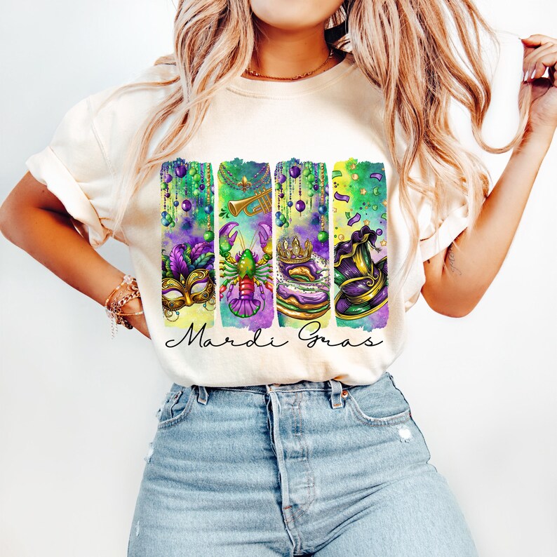 May include: A white t-shirt with a colorful graphic design featuring Mardi Gras symbols. The design includes a mask, a crab, a king cake, and a jester's hat. The text "Mardi Gras" is written below the design.