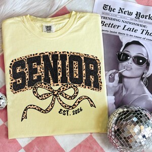 May include: A light yellow t-shirt with the word "SENIOR" in black glitter and leopard print, with a leopard print bow and the text "EST. 2026". A newspaper with the headline "Better Late Tha" is in the background.