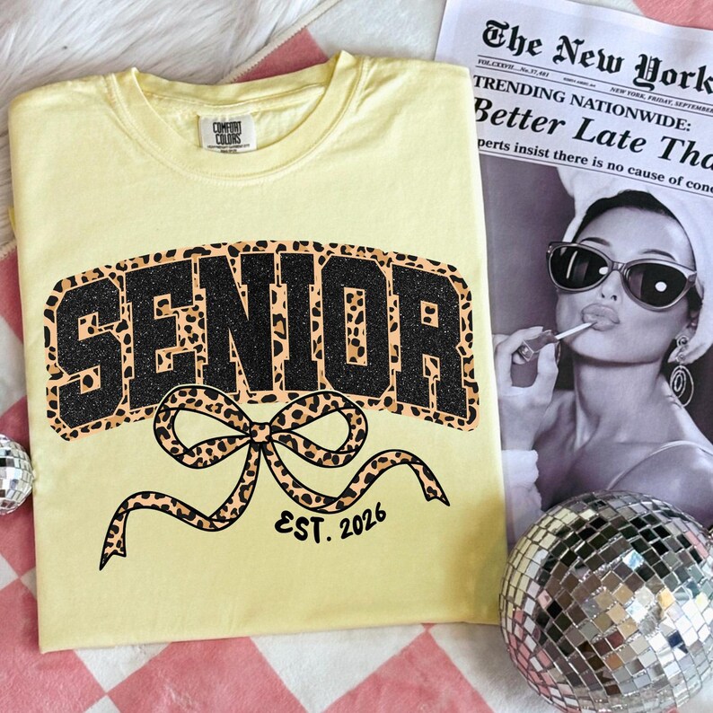 May include: A light yellow t-shirt with the word "SENIOR" in black glitter and leopard print, with a leopard print bow and the text "EST. 2026". A newspaper with the headline "Better Late Tha" is in the background.