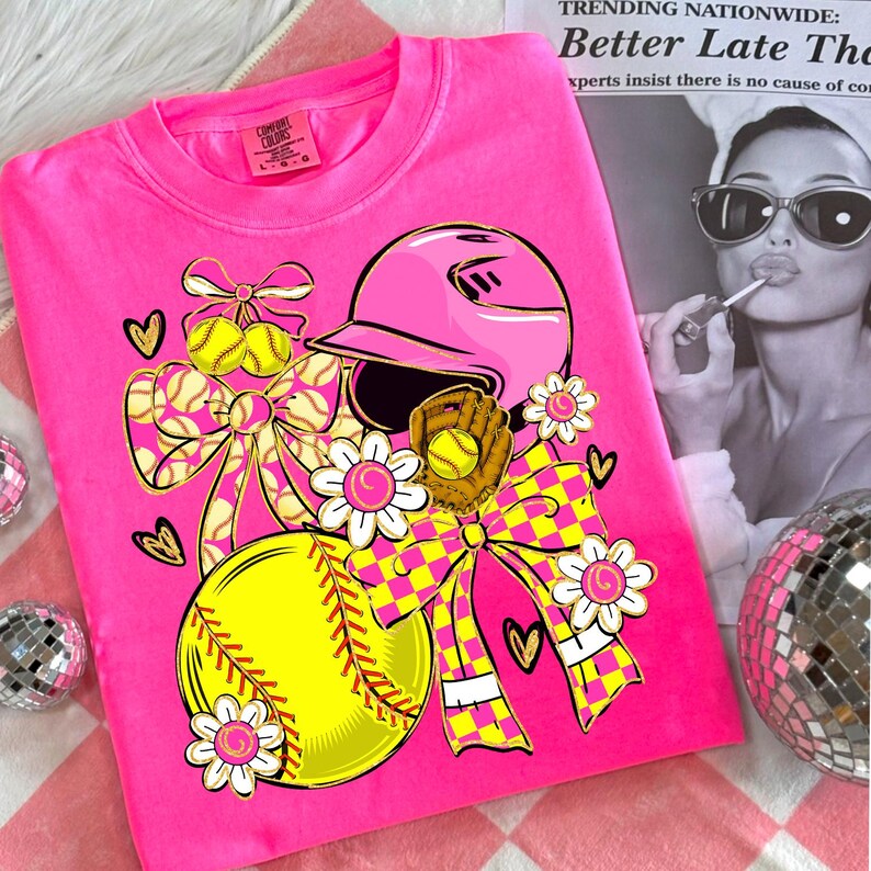 May include: A bright pink t-shirt featuring a softball design. The graphic includes a pink helmet, a softball, a glove, bows, and flowers. The text "COMFORT COLORS" is visible on the shirt's tag.