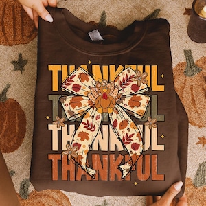 May include: A brown sweatshirt with the word "THANKFUL" in layered orange and yellow text. A large bow with fall leaves and a turkey graphic is centered on the shirt. The sweatshirt is held by hands.