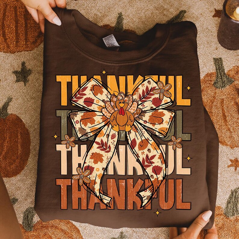 May include: A brown sweatshirt with the word "THANKFUL" in layered orange and yellow text. A large bow with fall leaves and a turkey graphic is centered on the shirt. The sweatshirt is held by hands.