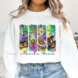 May include: White sweatshirt with a colorful graphic design featuring Mardi Gras themed imagery, including a crown, a mask, a lobster, a trumpet, and beads. The text "Mardi Gras" is printed below the graphic.