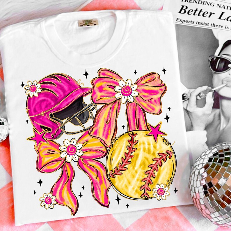 May include: White t-shirt featuring a softball design. The design includes a pink softball helmet, a yellow softball with red stitching, and two large pink and yellow bows. The design is accented with white flowers and pink stars.
