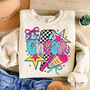Op de afbeelding: Cr&egrave;mekleurige sweatshirt met het woord "Cheer" in een kleurrijk, grafisch ontwerp. Het ontwerp bevat sterren, een megafoon en pompons in roze, blauw, geel en zwart. De sweatshirt ligt plat op een houten oppervlak.