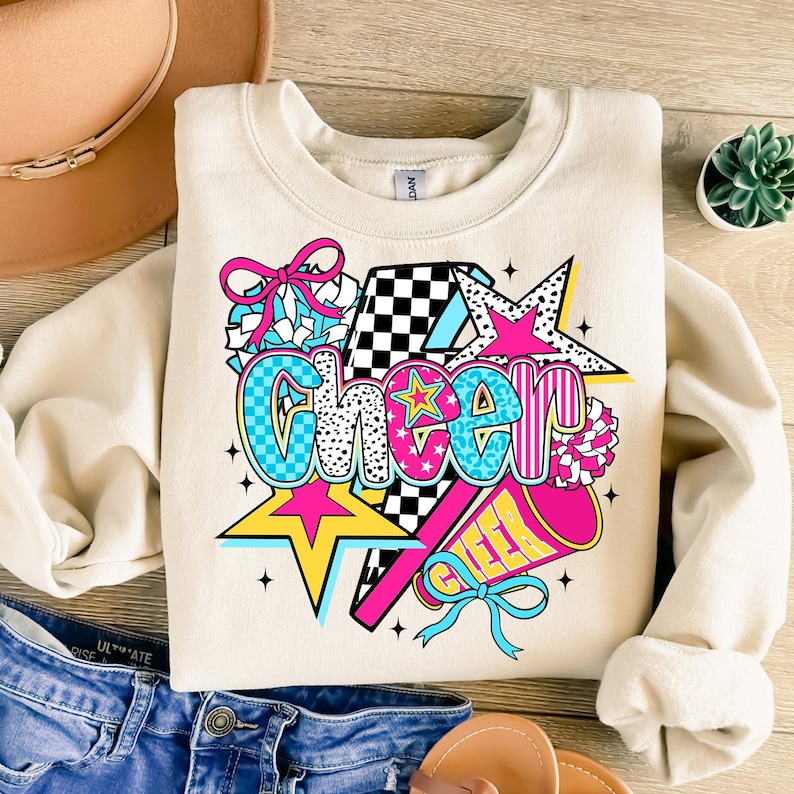 Op de afbeelding: Cr&egrave;mekleurige sweatshirt met het woord "Cheer" in een kleurrijk, grafisch ontwerp. Het ontwerp bevat sterren, een megafoon en pompons in roze, blauw, geel en zwart. De sweatshirt ligt plat op een houten oppervlak.