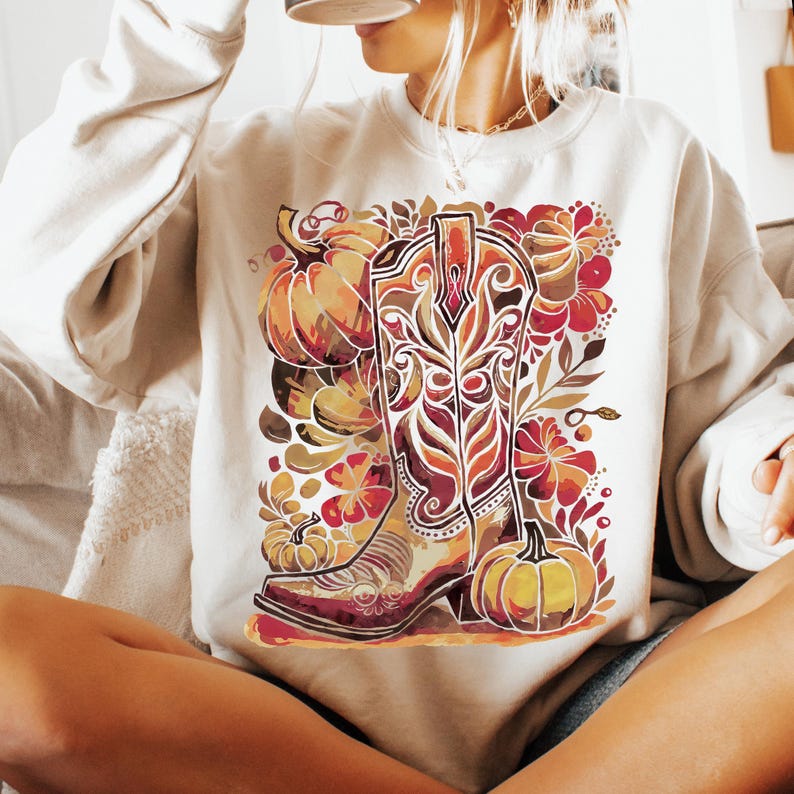 Op de afbeelding: Cr&egrave;mekleurige sweatshirt met een herfstthema. Het ontwerp bevat een gedetailleerde cowboylaars, pompoenen en bloemelementen in tinten oranje, rood en bruin. De algehele esthetiek is herfstig en rustiek.