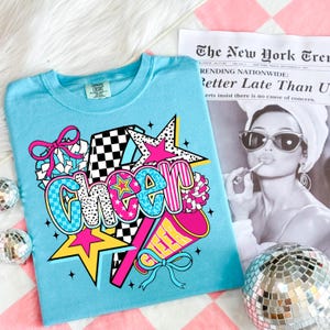 Op de afbeelding: Lichtblauw T-shirt met een kleurrijke "Cheer" afbeelding met sterren, een megafoon en strikken. Het shirt ligt op een roze en wit geruit oppervlak, met een krant op de achtergrond.