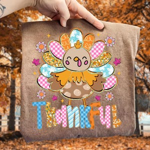 May include: A brown t-shirt featuring a colorful turkey design with the word "Thankful" in a playful font. The turkey has a patterned body and tail feathers in shades of pink, blue, orange, and yellow. Small flowers and leaves surround the turkey.