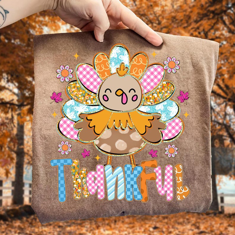 May include: A brown t-shirt featuring a colorful turkey design with the word "Thankful" in a playful font. The turkey has a patterned body and tail feathers in shades of pink, blue, orange, and yellow. Small flowers and leaves surround the turkey.