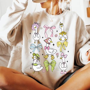 Puede incluir: Una sudadera blanca con un lindo dise&ntilde;o de dibujos animados de gansos con sombreros y lazos. Los gansos est&aacute;n rodeados de flores y lazos de varios colores.