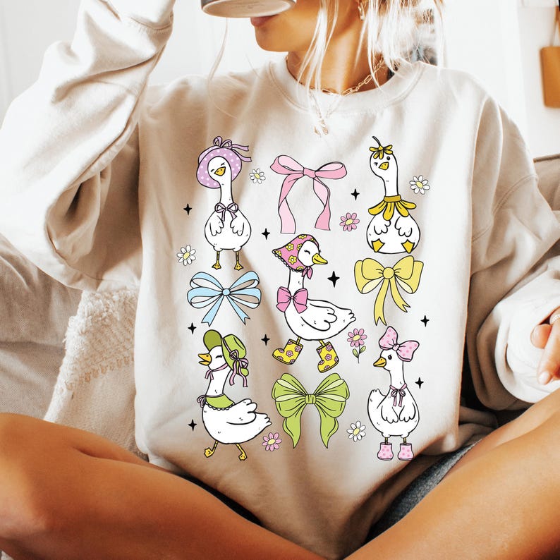 Puede incluir: Una sudadera blanca con un lindo dise&ntilde;o de dibujos animados de gansos con sombreros y lazos. Los gansos est&aacute;n rodeados de flores y lazos de varios colores.