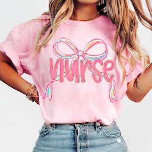 Pode incluir: Camiseta rosa claro com a palavra "Nurse" em uma fonte colorida e delineada, sob um design de la&ccedil;o correspondente. A camiseta &eacute; feita de um material macio e &eacute; adequada para uso casual.