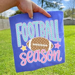 Może przedstawiać: Fioletowy t-shirt z napisem FOOTBALL w jasnoniebieskim kolorze w białe kropki, brązowo-białą grafiką piłki do futbolu amerykańskiego pośrodku oraz słowem season napisanym różową kursywą. Obok piłki znajdują się żółta i różowa gwiazdka.