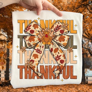 May include: A cream-colored t-shirt featuring the word "THANKFUL" in layered orange, brown, and tan lettering. A large bow with fall leaves and a turkey graphic is in the center. Small flower graphics and stars accent the design.