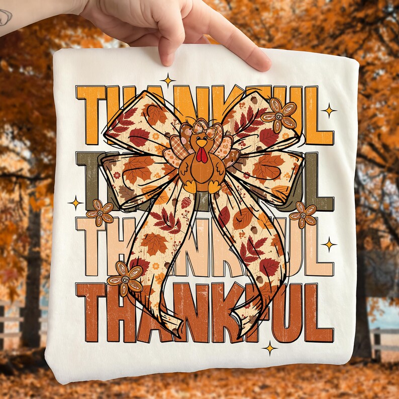 May include: A cream-colored t-shirt featuring the word "THANKFUL" in layered orange, brown, and tan lettering. A large bow with fall leaves and a turkey graphic is in the center. Small flower graphics and stars accent the design.