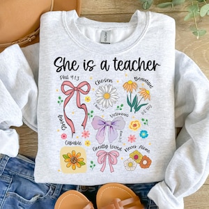 Op de afbeelding: Een lichtgrijs sweatshirt met de tekst "She is a teacher" in het zwart. Het ontwerp bevat bloemen- en strikgraphics met woorden als "Chosen", "Beautiful" en "Victorious".