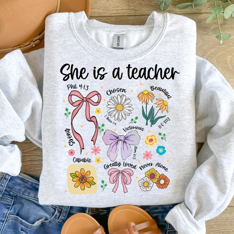Op de afbeelding: Een lichtgrijs sweatshirt met de tekst "She is a teacher" in het zwart. Het ontwerp bevat bloemen- en strikgraphics met woorden als "Chosen", "Beautiful" en "Victorious".
