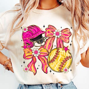 May include: A cream-colored t-shirt featuring a softball theme. The design includes a pink softball helmet, a yellow softball with red stitching, and pink and yellow bows adorned with white flowers. Black star accents complete the design.