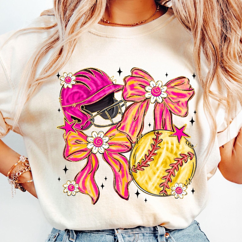 May include: A cream-colored t-shirt featuring a softball theme. The design includes a pink softball helmet, a yellow softball with red stitching, and pink and yellow bows adorned with white flowers. Black star accents complete the design.