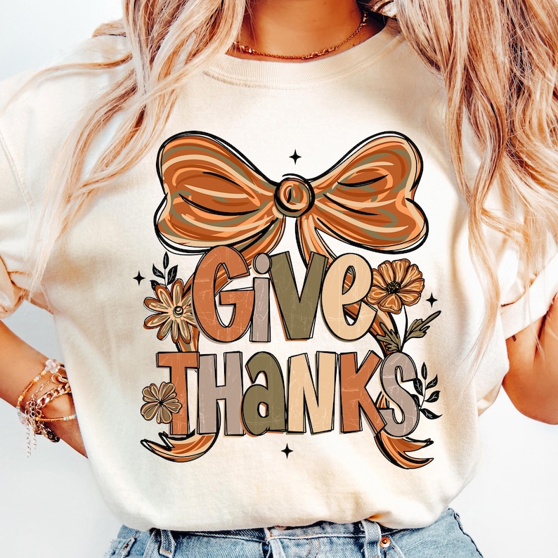 May include: Cream-colored t-shirt featuring a large bow design in shades of brown and orange. The words "GIVE THANKS" are printed in a mix of colors and fonts, with floral accents. A festive design for the Thanksgiving holiday.