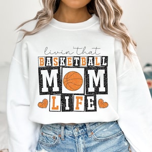 May include: White sweatshirt with a graphic print that says "Livin' that Basketball Mom Life" with a basketball graphic in the center.