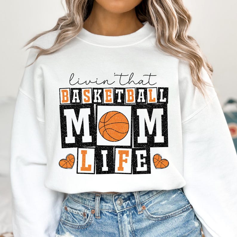 May include: White sweatshirt with a graphic print that says "Livin' that Basketball Mom Life" with a basketball graphic in the center.