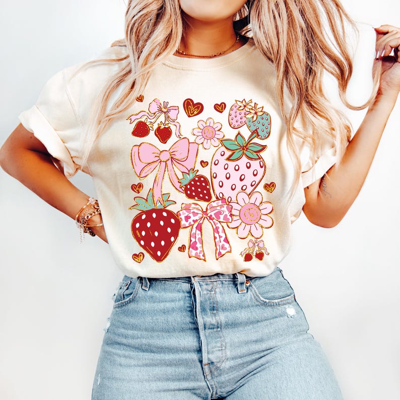 Op de afbeelding: Een cr&egrave;mekleurig T-shirt met een grafische print van aardbeien, strikken en bloemen in roze, rood, groen en goud.