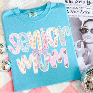 Puede incluir: Una camiseta azul claro con las palabras "Senior Mom" en un dise&ntilde;o de patchwork. Las letras est&aacute;n decoradas con telas florales y estampadas, y un lazo rosa acent&uacute;a el dise&ntilde;o. La camiseta est&aacute; sobre una superficie a cuadros rosa y blanco.