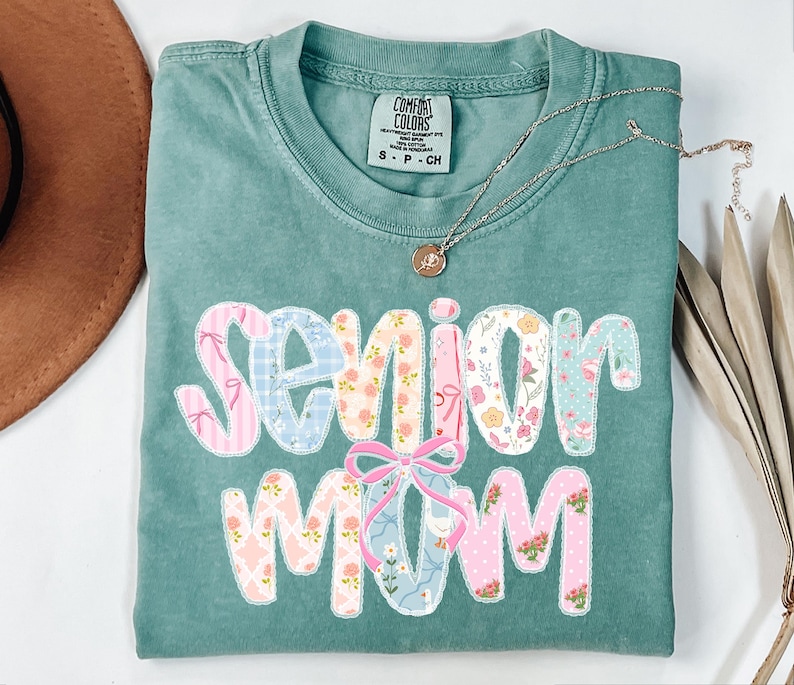 Puede incluir: Una camiseta Comfort Colors verde azulado con las palabras "Senior Mom" en un dise&ntilde;o de patchwork. Las letras est&aacute;n decoradas con patrones florales y lazos. Un collar plateado con un colgante est&aacute; sobre la camisa.