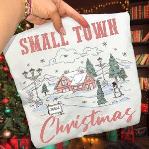 Może przedstawiać: Biała poduszka z ilustracją zimowej scenerii. Projekt przedstawia różowy dom, ośnieżone drzewa, bałwana oraz różowy napis "SMALL TOWN Christmas". Znak wyświetla numer 26554.