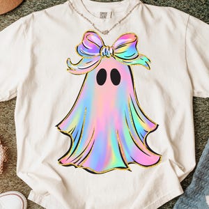 May include: Cream-colored t-shirt featuring a ghost design with a holographic, pastel color scheme. The ghost has a large bow and gold glitter accents. The shirt has the text "Comfort Colors" on the upper back.